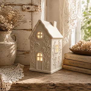 Könnte beinhalten: Eine cremefarbene, hausförmige Keramiklaterne mit geprägten Blumenmustern auf der Außenseite. Das Gebäude verfügt über eine mittige Bogentür, ein rundes Fenster darüber und ein Seitenfenster, die alle ein warmes Innenlicht ausstrahlen.
