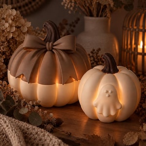Puede incluir: Dos decoraciones iluminadas con forma de calabaza. La calabaza más grande es beige con un tallo marrón oscuro y un lazo marrón. La más pequeña es blanca con un tallo marrón oscuro y una figura de fantasma en relieve. Ambas emiten luz cálida.