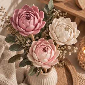 May include: A floral arrangement featuring three large, sculpted lotus-like flowers in shades of dusty rose, pale pink, and cream. The flowers are mounted on wooden stems and interspersed with dark green leaves and small, dried filler sprigs in a ribbed ceramic vase.