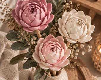 May include: A floral arrangement featuring three large, sculpted lotus-like flowers in shades of dusty rose, pale pink, and cream. The flowers are mounted on wooden stems and interspersed with dark green leaves and small, dried filler sprigs in a ribbed ceramic vase.