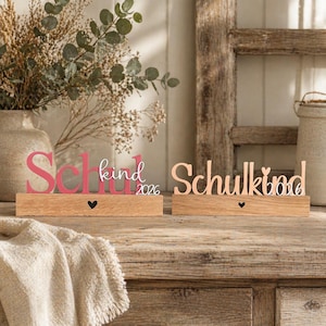 May include: Two wooden signs with the words "Schulkind" and "Schulkind 2026" in pink and peach lettering. Each sign sits on a rectangular wooden base with a small black heart. The signs are decorative.
