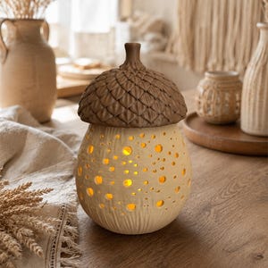 May include: A ceramic acorn-shaped lamp with a textured brown cap and a cream-colored base featuring numerous circular cutouts that emit a warm yellow light. The base has a subtle vertical ribbed pattern near the bottom, creating a glowing effect.