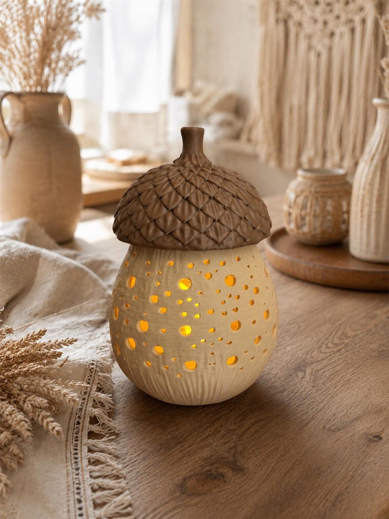 May include: A ceramic acorn-shaped lamp with a textured brown cap and a cream-colored base featuring numerous circular cutouts that emit a warm yellow light. The base has a subtle vertical ribbed pattern near the bottom, creating a glowing effect.