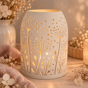 Può includere: Un portacandele cilindrico in ceramica bianca caratterizzato da intricati disegni ritagliati di fiori simili a denti di leone e punti sparsi. La struttura è illuminata dall'interno, proiettando una luce calda attraverso i motivi floreali.