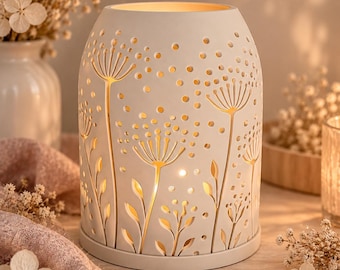 May include: A cylindrical white ceramic candle holder featuring intricate cut-out designs of dandelion-like flowers and scattered dots. The structure is illuminated from within, casting a warm glow through the floral patterns and circular perforations.