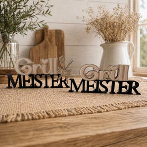 Può includere: Due insegne decorative con le parole "Grill Meister" in lettere nere e beige. Un'insegna ha uno striscione con la scritta "Papa" con utensili da grill. Le insegne sono su una superficie marrone.