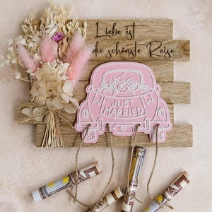 Könnte beinhalten: Ein Holzschild mit einem rosa Auto-Ausschnitt, der mit "JUST MARRIED" beschriftet ist, und einem Trockenblumenstrauß. Darüber ist der Text "Liebe ist die schönste Reise" eingraviert. Vier gerollte Papierröllchen hängen an Schnüren herab.