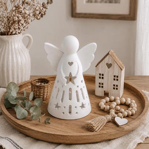Könnte beinhalten: Eine weiße Keramik-Engelsfigur mit ausgestanzten Stern- und Herzformen, platziert auf einem runden Holztablett neben einem kleinen Holzhaus-Ornament, einer Holzperlenkette mit Quaste und einem kleinen geflochtenen Korbbehälter.