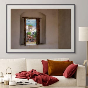 Puede incluir: Una impresión enmarcada de una ventana con vistas a una plaza. La ventana está enmarcada en madera oscura y empotrada en una pared de color claro. La escena fuera de la ventana muestra edificios, árboles y personas. Un sofá color crema con cojines rojos y naranjas está en primer plano.