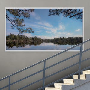 Puede incluir: Fotografía enmarcada de un paisaje con un lago sereno. La imagen captura un lago tranquilo que refleja el cielo y los árboles circundantes. El cielo es una mezcla de azul y blanco, con ramas de árboles que enmarcan las esquinas superiores.