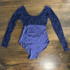 Puede incluir: Un maillot azul marino con mangas largas. La parte superior del maillot presenta un diseño de encaje, mientras que la parte inferior es de tela lisa y suave. Las mangas también son de encaje.