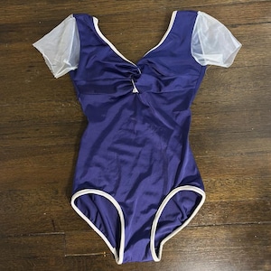 Puede incluir: Un maillot azul oscuro con ribete blanco y mangas blancas transparentes. El maillot tiene cuello en V y un detalle fruncido en el busto. La prenda está diseñada para danza o gimnasia.