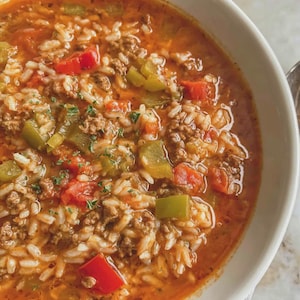 May include: A close-up shot of a bowl filled with a hearty soup. The soup features a rich, orange broth with visible ingredients like white rice, ground meat, red and green bell peppers, and herbs. The bowl is white, and the soup is steaming.