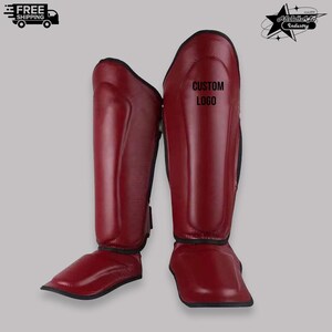 May include: Pair of burgundy-colored shin guards with black trim. The shin guards have a smooth, glossy finish and are designed to protect the lower legs. The text "CUSTOM LOGO" is printed on one of the guards. Ideal for martial arts or combat sports.