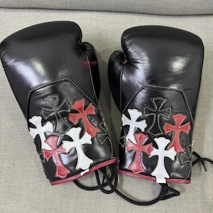 May include: Pair of black boxing gloves with decorative cross patterns in red and white. The gloves are made of leather and have a lace-up closure. The design features a red trim.