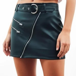 May include: Dark green faux leather skirt with a diagonal zipper and two smaller zippers. The skirt has a belt with a silver buckle. The skirt is a mini skirt.