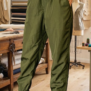 May include: Olive green nylon joggers with an elastic waistband and ankle cuffs. The pants are paired with yellow and white sneakers. The person is standing in a workshop with sewing supplies and fabric bolts.