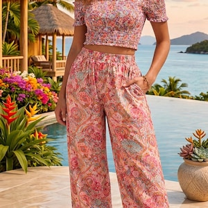 Puede incluir: Un conjunto de dos piezas con estampado floral. El top corto con hombros descubiertos y los pantalones de pernera ancha presentan un estampado de cachemira rosa, rojo y azul. El conjunto se lleva cerca de una piscina con un fondo tropical.
