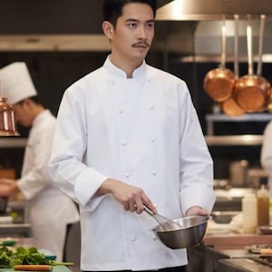 Puede incluir: Un chef con una chaqueta blanca de doble botonadura y pantalones negros, sosteniendo un batidor y un cuenco de metal. El chef está en una cocina comercial con ollas de cobre colgando en el fondo.