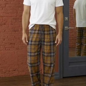 May include: A person wearing a white crew neck t-shirt and plaid pajama pants. The pajama pants have a brown, yellow, and navy blue plaid pattern. The person is standing in front of a brick wall and a mirror.