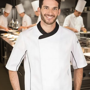 Puede incluir: Chaqueta de chef blanca con ribete negro y cuello negro. La chaqueta la lleva una persona sonriente en una cocina comercial. Otros chefs con uniformes y sombreros blancos son visibles en el fondo, trabajando en una encimera de acero inoxidable.