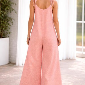 Puede incluir: Mono rosa claro con lunares blancos. El mono presenta un diseño de pernera ancha y tirantes anudados. La modelo lleva tacones beige. El mono está hecho de una tela ligera.