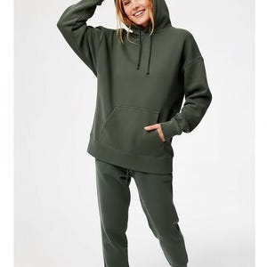 Può includere: Un completo coordinato composto da felpa con cappuccio e pantaloni della tuta verde oliva. La felpa con cappuccio ha un cappuccio con coulisse e una tasca frontale. I pantaloni della tuta hanno la vita con coulisse. L'outfit è realizzato in materiale morbido e confortevole.