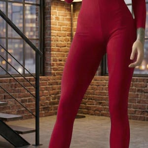 May include: A full-body, form-fitting red jumpsuit with a front zipper. The model is wearing dark blue boots. The background features a brick wall and large windows.