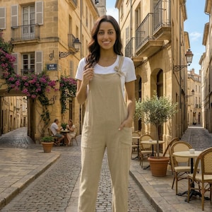 Puede incluir: Una mujer con un mono beige y una camiseta blanca en una calle europea. El mono tiene bolsillos y un lazo decorativo en los tirantes. La calle adoquinada está bordeada de edificios y cafés al aire libre.