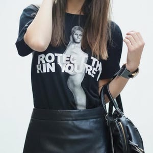 Puede incluir: Camiseta negra con el texto "PROTECT THE SKIN YOU'RE IN" y un gráfico de una mujer. El conjunto incluye una falda de cuero negra con tachuelas plateadas, gafas de sol negras, una mochila negra y un reloj.
