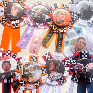 May include: A collection of colorful rosette badges featuring Formula 1 drivers. Each badge has a central image of a driver, surrounded by ribbons in various colors, including orange, black, white, and blue. Some ribbons have checkered patterns and star accents.
