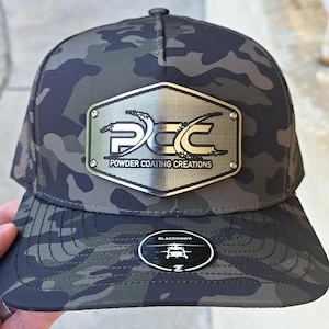 Pode incluir: Boné de beisebol camuflado com um emblema metálico. O emblema apresenta o texto "PCC Powder Coating Creations". O boné tem uma aba curva e um rótulo Blackhawk.