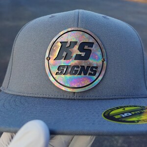 May include: Grey fitted baseball cap with a flat peak. The cap has a circular emblem with the text "KS SIGNS" in black. The emblem has an iridescent, oil-slick effect. A yellow and green sticker is on the peak.