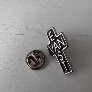 Può includere: Una spilla da bavero color argento a forma di croce con la scritta "EVANST" in nero. La spilla ha un bordo nero e un retro argentato con una chiusura.