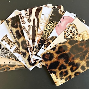 Puede incluir: Un conjunto de recambios decorativos para planificador con varios diseños de estampados de animales, incluyendo leopardo y cebra, así como motivos florales. Los recambios están dispuestos en forma de abanico, mostrando los diferentes diseños y texturas. Cada recambio tiene agujeros preperforados.