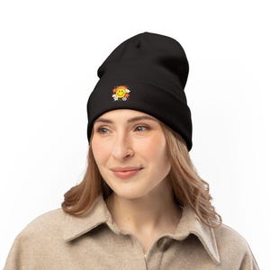 May include: Black beanie hat with a colourful embroidered patch showing a smiling face, rainbow, and flowers. The hat is made of a soft material and is designed to be worn on the head. The patch is centered on the front of the beanie.