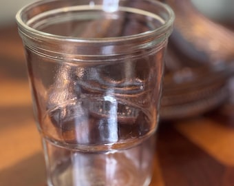 May include: A clear, empty glass jar with a wide mouth and a slightly tapered shape. The jar has a thick rim and a distinct horizontal line near the middle. The background features a blurred wooden object and a wooden table.