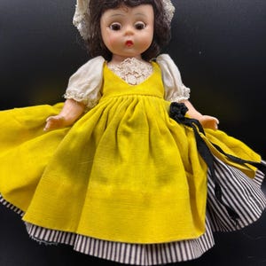 May include: A vintage doll with brown hair and a white lace bonnet. The doll wears a yellow dress with white sleeves and a black and white striped skirt. The doll has a painted face with red lips and is posed against a black background.