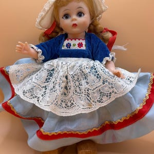 May include: A vintage doll wearing a blue velvet top with floral embroidery, a white lace skirt, and a light blue skirt with red trim. The doll has blonde hair, blue eyes, and a white bonnet with red ribbons. The doll is standing on two wooden blocks.