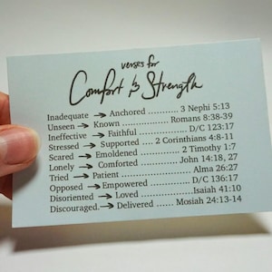 May include: A light blue card with the words "verses for Comfort & Strength" in script. The card lists pairs of words such as "Inadequate" to "Anchored" with corresponding Bible verses. The card is held by a hand.