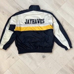 May include: A navy blue and white jacket with a yellow stripe across the chest. The word "JAYHAWKS" is embroidered in black on the back. The jacket has a ribbed hem and cuffs.