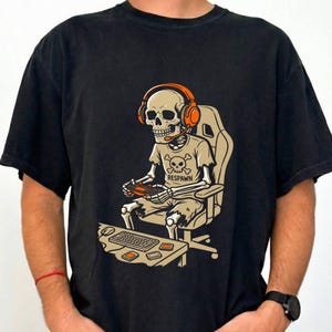 May include: A black t-shirt featuring a graphic of a skeleton wearing orange headphones and a t-shirt with a skull and crossbones design. The skeleton is sitting in a gaming chair, holding a controller, with a keyboard on a desk in front.