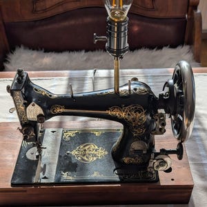 Könnte beinhalten: Eine antike, schwarze Nähmaschine mit goldenen Verzierungen, die zu einer Lampe umfunktioniert wurde. Die Lampe hat eine Edison-Glühbirne und steht auf einem Holzsockel, was ein einzigartiges Dekorationsobjekt ergibt.