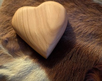 Op de afbeelding: Een houten hartvormig object met een lichte bruine kleur en zichtbare houtnerf. Het hart ligt op een oppervlak met een bruin en wit patroon, wat een contrast creëert. Het is waarschijnlijk een decoratief item of een klein beeldhouwwerk.