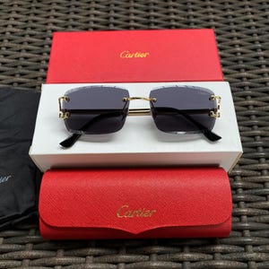 May include: Square-framed sunglasses with dark lenses and gold-colored accents. The sunglasses are displayed with a red box and case, both with the brand name "Cartier" in gold lettering. The lenses are a dark gray color.