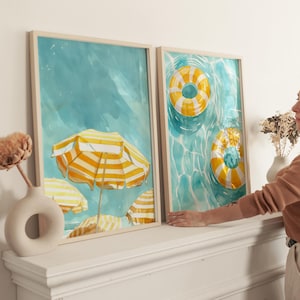 May include: Two framed watercolor prints. One features yellow and white striped beach umbrellas, the other shows yellow and white striped pool floats in a turquoise pool. The frames are light wood.