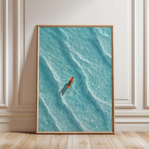 May include: A framed print featuring an aerial view of a person on an orange surfboard in clear turquoise water. The water's surface has gentle waves and sunlight reflections. The frame is a light brown color.