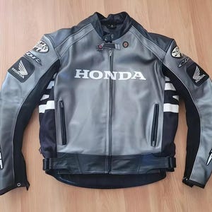 Pode incluir: Casaco de motociclista cinza e preto com a palavra "HONDA" no peito. O casaco tem listras brancas nas mangas e a palavra "ROCKET" impressa na manga esquerda. Fecho de correr e vários logotipos.
