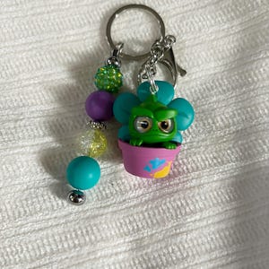May include: A whimsical keychain featuring a green creature in a pink flower pot. The keychain has a silver ring and clasp, with a beaded strand of green, purple, yellow, and teal beads. A fun accessory.