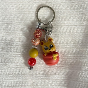 May include: A keychain featuring a yellow bear figure in a red pot. The keychain has a silver ring and chain, with a beaded strand of red, yellow, and orange beads. The beads are accented with silver beads.